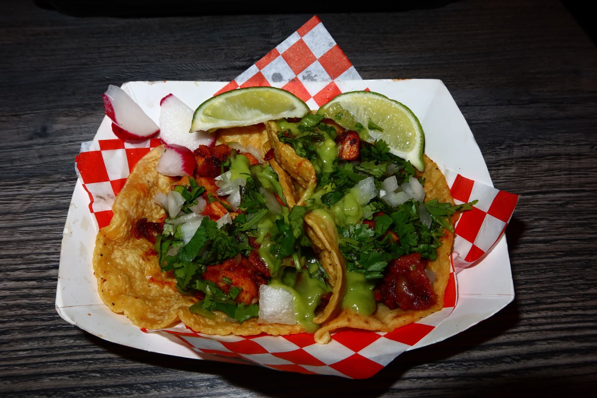 A paper tray of three tacos on red-and-white checkered paper, topped with cilantro, diced onion, and a green sauce, with lime wedges and sliced radishes